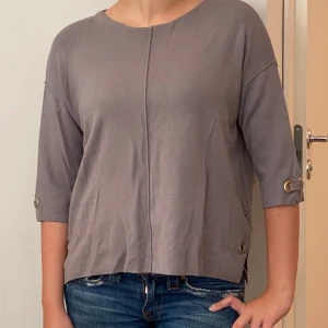 Grå tröja från Rabens Saloner - Säljer en stilren grå tröja från Rabens Saloner i storlek XS/S. Tröjan har trekvartsärmar och dekorativa knappar på ärmarna och sidorna. Perfekt för en avslappnad vardagslook. Materialet känns mjukt och bekvämt. Passar bra till jeans eller kjol!