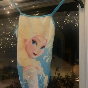 Blå klänning med Disney-motiv - Säljer en superfin blå baddräkt med motiv av en karaktär från Disney. Baddräkten har smala axelband och är perfekt för alla små fans av Disney. Den är i bra skick och redo för nya äventyr!