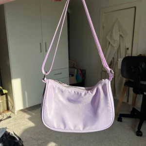 Lila väska - Säljer en väska i en härlig ljuslila färg! Bandet är justerbart så man kan ha den antingen som en mini-axelväska eller cross body bag. Finns en liten grå fläck på vardera sida men det var svårt att fota. Annars bra skick!💞 