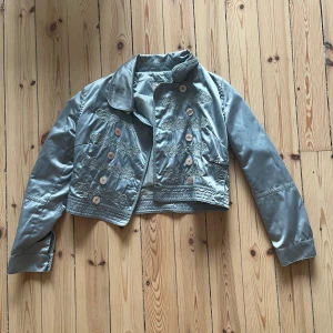 Ljusblå jacka med broderade detaljer - Säljer en superfin ljusblå jacka med snygga broderade detaljer. Den har en kort, croppad stil och är dekorerad med knappar framtill. Jackan är i bra skick och redo för nya äventyr! 