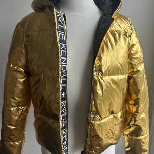 Guldig pufferjacka från Kendall + Kylie - Säljer en supercool guldig pufferjacka från Kendall + Kylie för OVS. Jackan har en glansig finish och är perfekt för att sticka ut i mängden. Den har en dragkedja framtill och en mysig huva för extra värme. Perfekt för vintern och hösten när du vill hålla dig varm och stilren.