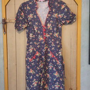 Blommig klänning från Trollied Dolly - Säljer en superfin blommig vintage klänning från Trollied Dolly i storlek S. Klänningen har en mörkblå bas med ett sött mönster av små vita prickar och smultronblommor. Den har korta ärmar och en v-ringad hals med röda knappar som detalj. 