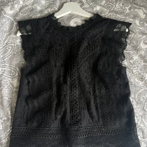 Svart spetsblus från ONLY - Säljer en elegant svart spetsblus från ONLY i storlek XS. Blusen har ett vackert spetsmönster och är ärmlös med en dragkedja i ryggen. Perfekt för festliga tillfällen eller en utekväll. Den är både stilren och chic!