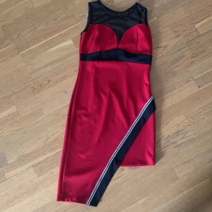 Röd ärmlös klänning från Jenny Pronto Moda - Säljer en snygg röd klänning från Jenny Pronto Moda. Den är ärmlös med en cool svart mesh-detalj upptill och ett svart band i midjan. Klänningen har en diagonal svart rand med vit kant som ger en modern touch. Perfekt för festliga tillfällen!