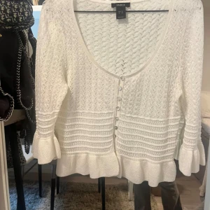 Vit stickad kofta från Uniqlo - Säljer en superfin vit stickad kofta från Uniqlo. Den har en v-ringad design med knappar framtill och långa ärmar. Koftan har en snygg volangdetalj vid ärmslut och nederkant som ger den en extra touch. Perfekt för både höst och vår!