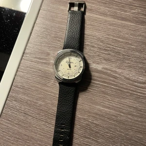 Svart klocka från Diesel - Säljer en snygg klocka från Diesel med svart läderarmband och en stilren urtavla. Klockan har en silverfärgad boett och tydliga markeringar på urtavlan. Pris går att diskuteras😊behövs nytt batteri då det nuvarande är slut