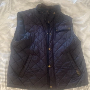 Blå quiltad väst från Polo Ralph Lauren - Säljer en snygg blå quiltad väst från Polo Ralph Lauren. Västen har en stilren design med knappar och dragkedja framtill. Perfekt för höst och vår när du vill hålla dig varm men ändå se cool ut. Den har praktiska fickor och en bekväm passform.
