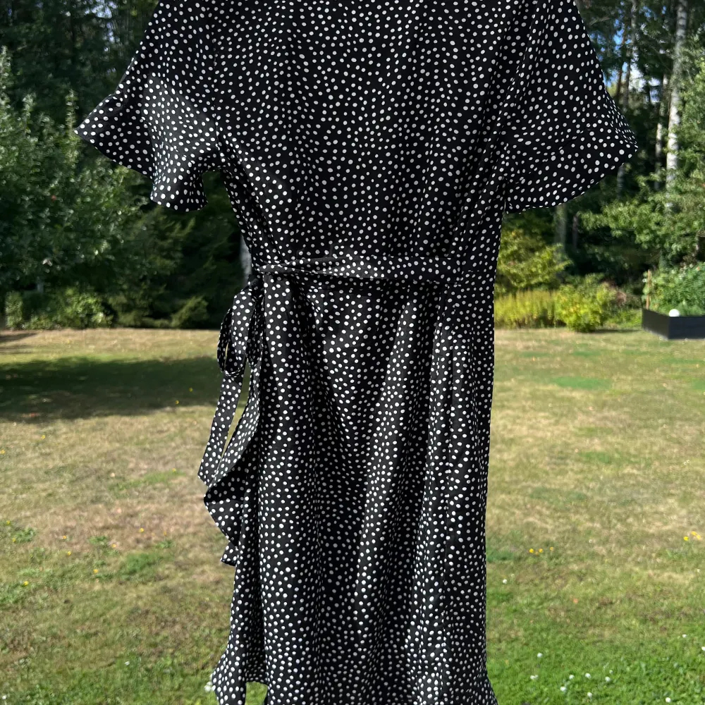 Svart knälång klänning med vita prickar. Klänningen läggs omlott och knyts i midjan på vänster sida. Volanger längst ner och i ärmsluten 🌸💞 (ALDRIG ANVÄND) Längd: 95 cm Axel: 35 cm. Mekot.