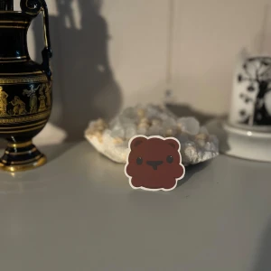 Söt björnringsdekal - En supergullig klistermärke i form av en björn med ett glatt ansikte. klistermärket har en brun färg med en regnbågseffekt som syns i ljuset. Perfekt för att dekorera dina saker och ge dem en personlig touch!