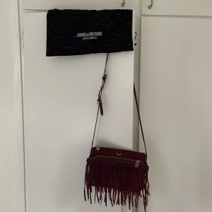 Vinröd Zadig & Voltaire väska med fransar - Zadig & Voltaire väska i mocka och läder som inte längre finns. Använd så är lite slitage på bandet (se bild) i övrigt bra skick. Bandet går att reglera och kan även bäras som clutch. Rymlig med tre fack och ett innerfack. Dustbag följer med.