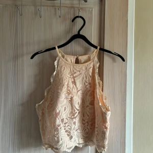 Beige spetslinne - Säljer ett superfint beige/ljusrosa spetslinne med vackra detaljer. Det är ärmlöst och har en luftig passform, perfekt för sommardagar eller festliga tillfällen. Linnet har en knäppning i nacken och är gjort i ett mjukt och bekvämt material.
