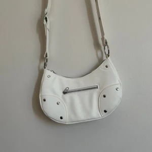 Vit axelväska från H&M Divided - Säljer en superfin vit axelväska från H&M Divided. Den har en justerbar axelrem och en praktisk dragkedja framtill. Väskan är dekorerad med nitar som ger den en cool look. Perfekt för både vardag och fest! Avänd endast en gång.