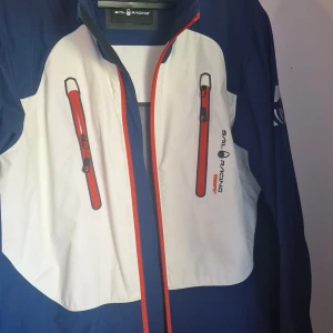 Blå och vit jacka från Sail Racing - Säljer en snygg blå och vit jacka från Sail Racing. Den har en cool design med röda dragkedjor och logotyp på bröstet. Perfekt för vår och höst med sin sportiga stil och funktionella detaljer.