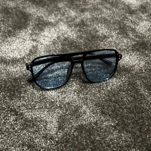 Svarta solglasögon med blå linser - Säljer ett par snygga svarta solglasögon med blå tonade linser. De har en klassisk aviator-stil med en tunn ram och dubbla bryggor. Perfekta för en cool look i sommar! 