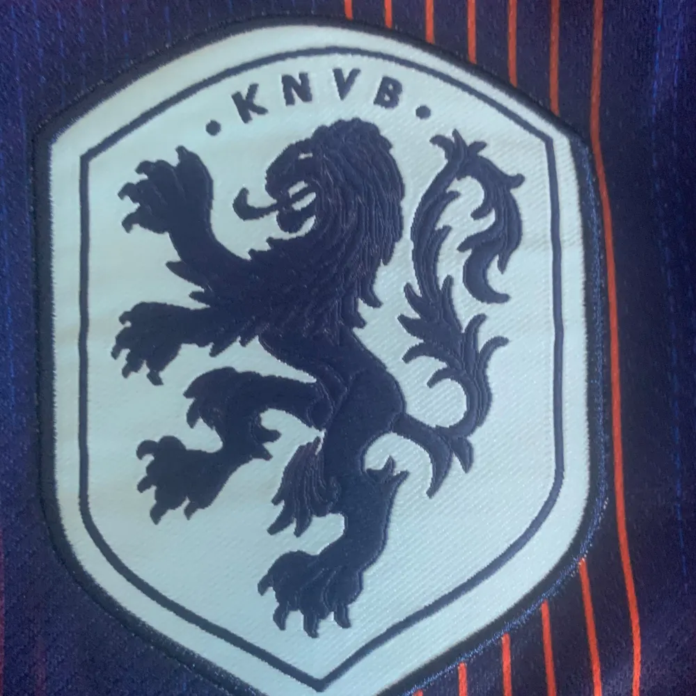 Säljer en blå fotbollströja från Nike med ett coolt mönster i orange och svart. Tröjan har korta ärmar och är gjord av 100% polyester, vilket gör den lätt och bekväm. Den har ett KNVB-märke på bröstet och en orange krage. Perfekt för fotbollsfans! Ingen på ryggen.. T-shirts.