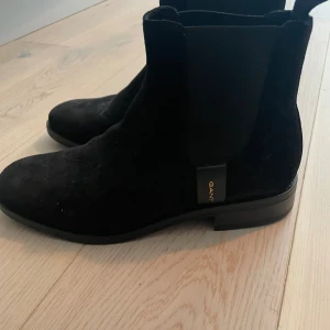 Svarta Chelsea boots från Gant - Säljer ett par stilrena svarta Chelsea boots från Gant. De är gjorda i mocka och har en klassisk design med elastiska paneler på sidorna för enkel på- och avtagning. Perfekta för både höst och vinter!