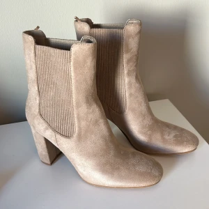 Beige stövletter i mocka - Säljer ett par snygga beige stövletter i mocka. De har en stilren design med en bekväm klack och elastiska paneler på sidorna för enkel på- och avtagning. Perfekta för både vardag och fest!