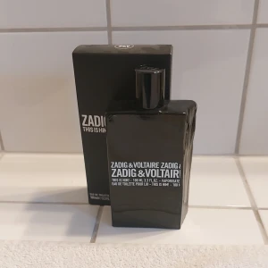 Zadig & Voltaire This is Him! Eau de Toilette - Säljer en cool och stilren parfymflaska från Zadig & Voltaire. Flaskan är svart och har en minimalistisk design med vit text. Det är en Eau de Toilette för herrar, perfekt för den som vill ha en modern och maskulin doft. Det är ca 90-93 ml kvar. 