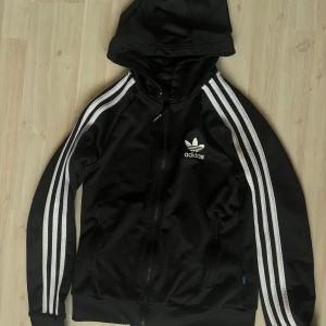 Adidas jacka - Svarta adidas jacka med luva och vita detaljer. Dragkedja längs med hela samt två fickor med dragkedja. Snörningen i luvan är inte kvar. Normal i storlek. 