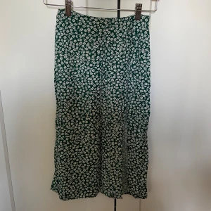 Grön blommig kjol från H&M Divided - Säljer en superfin grön kjol med vita blommor från H&M Divided. Kjolen är i storlek EUR 32 och har en söt knappdetalj och slits framtill. Perfekt för vår och sommar! 🌸
