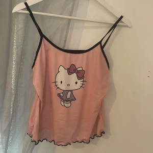 Rosa Hello Kitty linne - Söt rosa linne med Hello Kitty-tryck på framsidan. Linnet har smala svarta axelband och en vågig kant nedtill. Perfekt för en avslappnad stil.