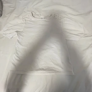 Vit t-shirt från Zara - Säljer en vit t-shirt från Zara med korta ärmar och dekorativa detaljer vid axlarna. Perfekt för en stilren och enkel look.