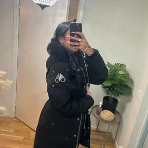 Svart parkajacka med pälsdetaljer - Snygg svart parkajacka från Moose Knuckles med pälsdetaljer på huvan. Jackan har en dragkedja och knappar framtill samt en logotyp på ärmen. Perfekt för kalla dagar.