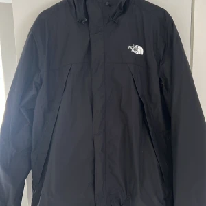 Svart regnjacka från The North Face - Säljer en svart regnjacka från The North Face med huva och dragkedja. Jackan har en enkel och stilren design med The North Face-logga på bröstet och ryggen. Perfekt för regniga dagar.