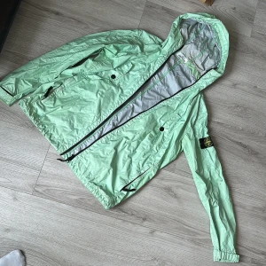 Mintgrön vindjacka från Stone Island - Snygg mintgrön vindjacka från Stone Island med huva och dragkedja. Jackan har långa ärmar och en stilren design med två fickor framtill. Perfekt för vår och sommar!