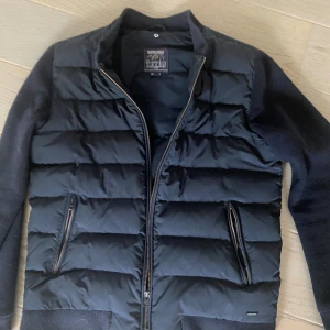 Svart pufferjacka från Woolrich - Säljer en svart pufferjacka från Woolrich med dragkedja och två fickor framtill. Jackan har långa ärmar och en stilren design, perfekt för kyligare dagar. Den är storlek L men passar perfekt M och killen på bilden är 183 och väger 68kg: ny pris 5000+ kr