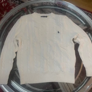 Vit stickad tröja från Polo Ralph Lauren - Säljer en stilren vit stickad tröja från Polo Ralph Lauren med klassiskt kabelstickat mönster. Tröjan har långa ärmar och en liten broderad logotyp på bröstet. Perfekt för en elegant och avslappnad look.