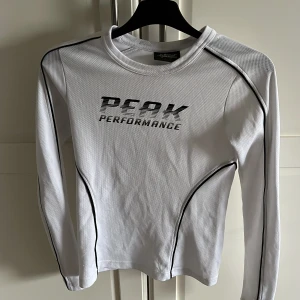 Vit långärmad tröja från Peak Performance - Säljer en vit långärmad tröja från Peak Performance med svart text och svarta detaljer längs ärmarna och sidorna. Bra att ha när man åker skidor.Tröjan har en tight passform och är tillverkad i ett syntetmaterial som andas. PRISET KAN DISKUTERAS