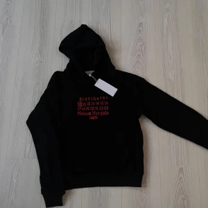 Maison Margiela Hoodie - Säljer en stilren och exklusiv Maison Margiela hoodie i svart med röd text. Tröjan har det ikoniska trycket, där number 10 är markerat, vilket symboliserar Maison Margielas herrkollektion.