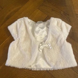 Vit pälsväst för barn - Söt vit pälsväst för barn från Lindex. Västen har en fin rosett framtill och är perfekt för att ge en extra touch till en outfit. Den är mjuk och har en glansig insida. Storlek 98/104