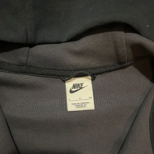 Svart och grå hoodie från Nike - Säljer en snygg svart och grå hoodie från Nike med dragkedja och huva. Jackan har en cool design med kontrasterande paneler och en ficka med dragkedja på ärmen. Perfekt för en sportig look!