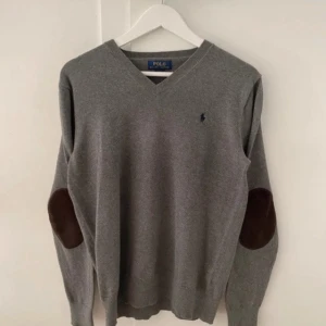 Grå tröja med bruna armbågslappar från Polo - Säljer en stilren grå tröja från Polo med bruna armbågslappar. Tröjan har en v-ringad design och är tillverkad i 100% bomull. Perfekt för en klassisk och avslappnad look.