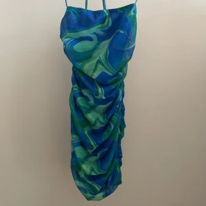 Blå och grön klänning med mönster - Snygg klänning i blått och grönt med ett abstrakt mönster. Den har smala axelband och en åtsittande passform som framhäver figuren. Perfekt för en somrig look.