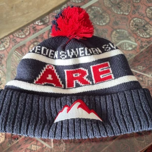 Blå och röd Åre mössa - Snygg stickad mössa i blått med röd och vit text 'ÅRE' och 'SWEDEN'. Mössan har en röd och blå tofs på toppen och ett broderat bergsmotiv framtill. Perfekt för kalla dagar med en chill outfit, priset ej hugget i sten!