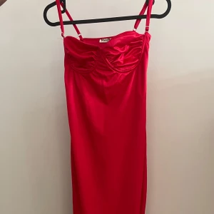 Röd satin axelbandslös klänning  - Snygg röd satin axelbandslös klänning med smala axelband och en elegant slits. Perfekt för en kväll ute eller en speciell tillställning. Klänningen har en figurnära passform och är tillverkad i ett mjukt material som ger en lyxig känsla.