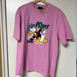 Rosa t-shirt med Kalle Anka-tryck - Säljer en cool rosa t-shirt från H&M med ett färgglatt Kalle Anka-tryck och texten 'Pow!'. Perfekt för dig som gillar serietidningar och vill ha en unik stil. T-shirten är kortärmad och gjord i mjukt bomullsmaterial.