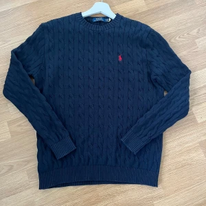 Mörkblå stickad tröja från Ralph Lauren - Säljer en stilren mörkblå stickad tröja från Ralph Lauren med kabelstickat mönster. Tröjan har en klassisk rund hals och långa ärmar. Den ikoniska röda logotypen är broderad på bröstet. Perfekt för en elegant och avslappnad look. Helt ny och sitter jättebra!