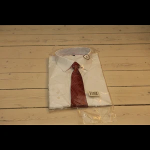 Vit skjorta med röd slips - Snygg vit skjorta med en stilren röd slips. Perfekt för formella tillfällen. Skjortan har en klassisk krage och är långärmad. Den är förpackad i plast och redo att användas.