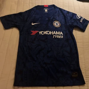 Blå Chelsea fotbollströja från Nike - Säljer en blå Chelsea fotbollströja från Nike med korta ärmar och snyggt mönster. Tröjan har klubbens emblem på bröstet och Yokohama Tyres som sponsorlogga. Perfekt för alla Chelsea-fans!