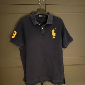 Mörkblå pikétröja från Ralph Lauren - Säljer en klassisk mörkblå pikétröja från Ralph Lauren med den ikoniska gula logotypen på bröstet. Tröjan har korta ärmar och en krage med knappar. Perfekt för en stilren look.