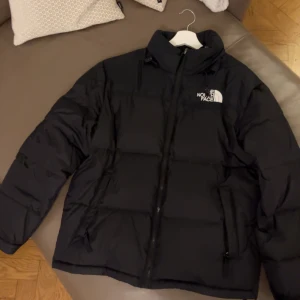 Svart dunjacka från The North Face - Säljer en svart dunjacka från The North Face. Perfekt för kyliga dagar med sin pufferdesign som håller värmen. Klassisk och stilren med logotyp på bröstet.
