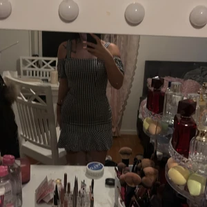 Svartvit rutig klänning - Säljer en söt svartvit rutig klänning med offshoulder-design och volangdetalj vid fållen. Klänningen har en tight passform som framhäver figuren och är perfekt för en somrig look. Den har smala axelband och en kort längd som ger en ungdomlig stil.