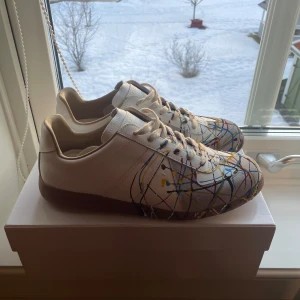 Vita sneakers från Maison Margiela - Unika vita sneakers från Maison Margiela med färgglatt stänkmönster i blått, rött, gult och svart. Skorna har snörning och är tillverkade i skinn. Perfekta för den som vill sticka ut med en konstnärlig touch. Helt nya skick 10/10