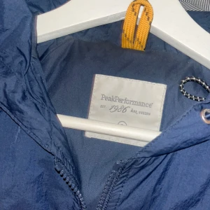 Blå vindjacka från Peak Performance - Snygg blå vindjacka från Peak Performance med dragkedja framtill. Jackan har en sportig look och är perfekt för blåsiga dagar. Den har flera praktiska fickor med dragkedjor och en justerbar huva.