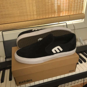 Svarta slip-on sneakers från Etnies - Säljer ett par helt nya svarta slip-on sneakers från Etnies som säljs pågrund av fel storlek.Med en vit sula och logotyp.Nypris 900kr. Skorna har en stilren design med en bekväm passform, perfekt för vardagsbruk. De är tillverkade i syntetmaterial och har en klassisk gummisula för bra grepp.