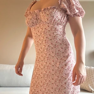 Blommig klänning från NA-KD strl 36 - Säljer en söt blommig klänning från NA-KD i storlek 36. Klänningen har puffiga kortärmar och en v-ringad design. Perfekt för sommarens alla tillfällen med sitt fina blommönster i rosa och beige toner. Oanvänd men upptvättad. Passar 36/38.